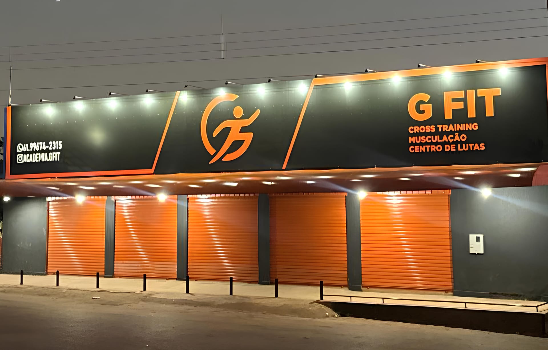 Fachada da GFIT Academia à noite, com a placa iluminada, o logotipo em laranja e as portas fechadas.